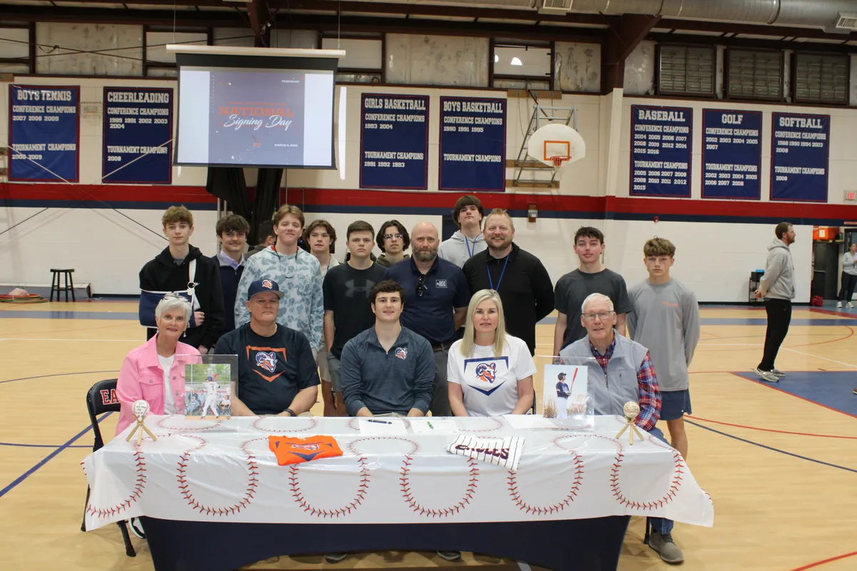 Group portrait in a gym: students and coaches stand behind a long table draped with a baseball-themed cloth for a signing day ceremony.